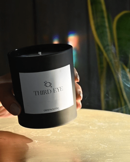 Black Third Eye Candle in a matte container with a white label, held in a person's hand on a wooden surface.