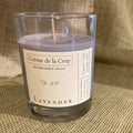 Lavender soy candle in a clear glass container with a beige label, placed on textured beige fabric.