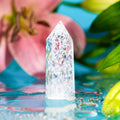 Limitless Manifestation - Ice Quartz Point with clear, faceted crystal on blue water surface, pink flower, and green leaves.
