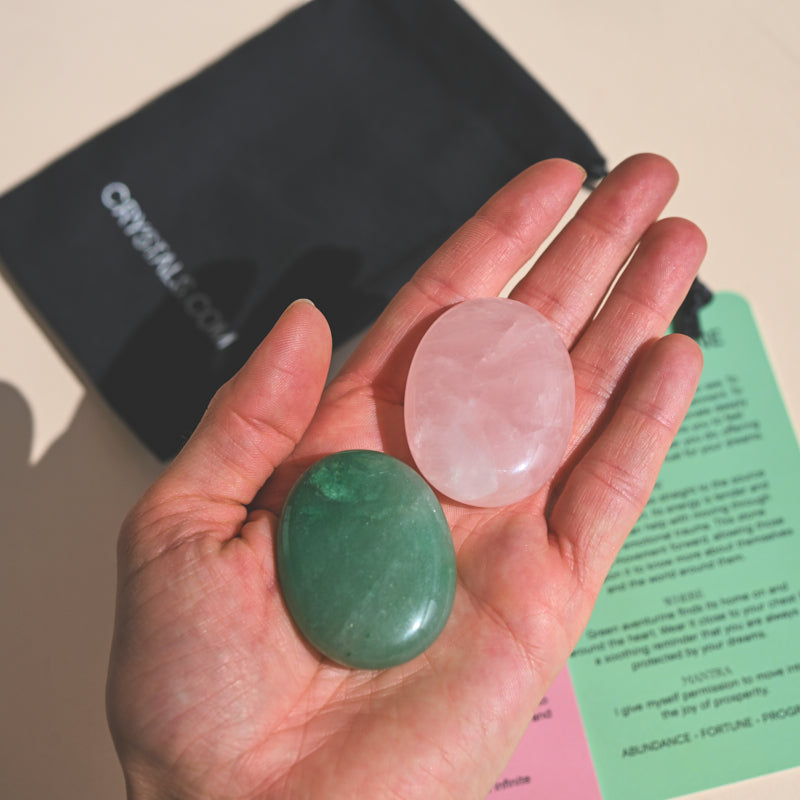 Green Aventurine Palm Stone held in hand with pink quartz palm stone, black pouch, and informational card in background.