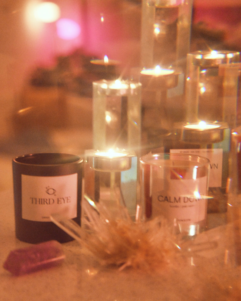 Third Eye Candle with lit flames in clear glass holders, black and white labels, surrounded by pink crystal and dried